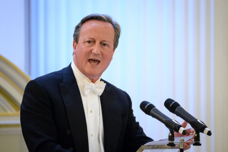 David Cameron smiling at a press conference podium, wearing a navy suit and blue tie, addressing the media in a formal set...