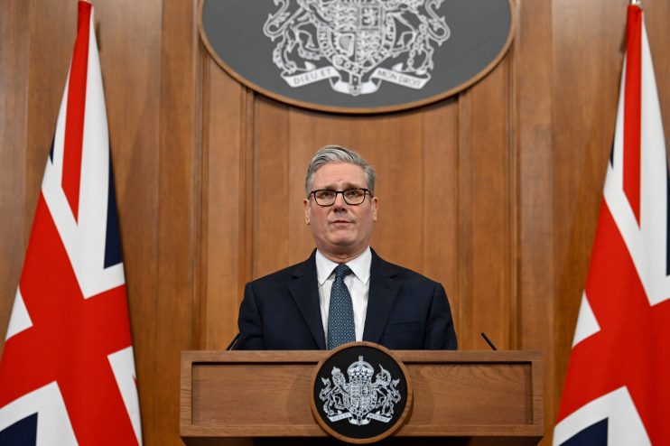 Keir Starmer discussing Iran policy at press conference podium with flags in the background