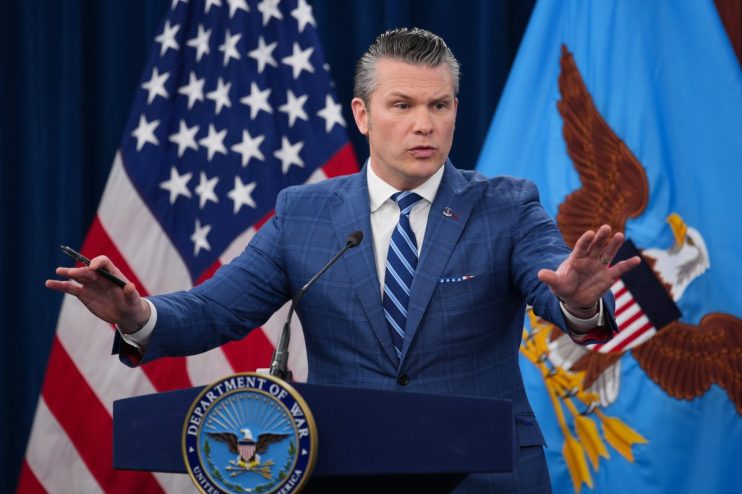 Pete Hegseth speaking passionately at a podium, wearing a suit, with an American flag backdrop and engaging audience.