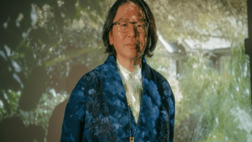 Person with long dark hair and glasses wearing a blue patterned jacket, projection of greenery behind them.