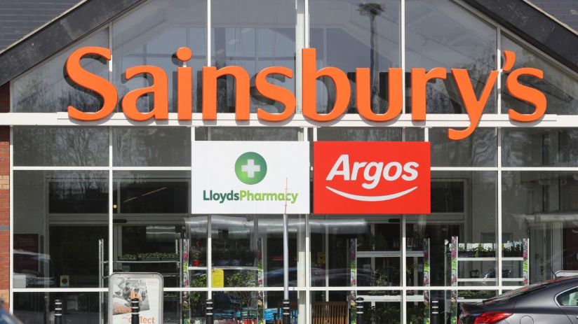 Sainsburys supermarket entrance with prominent Argos and Lloyds Pharmacy signs, reflecting the companys acquisitions.
