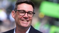 Andy Burnham, Mayor of Greater Manchester, smiling in glasses, dark jacket, light blue shirt, blurred green background.