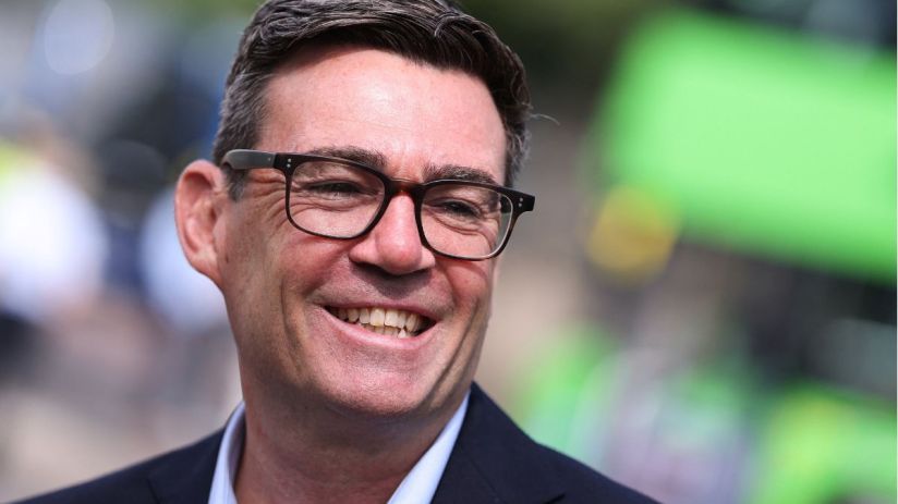 Andy Burnham, Mayor of Greater Manchester, smiling in glasses, dark jacket, light blue shirt, blurred green background.