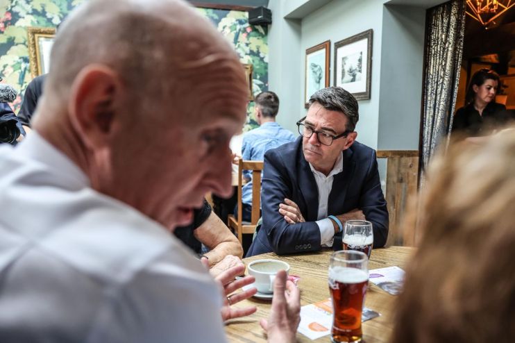 Andy Burnham, Mayor of Greater Manchester, in a dark suit and glasses, listening intently at a wooden table.