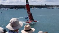 Red foiling sailboat racing on blue water with a bridge and city in the background, spectators watching