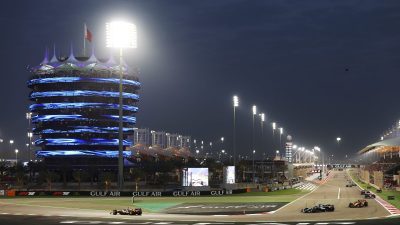 Night race at Bahrain International Circuit with brightly lit tower, grandstands, and F1 cars on track.