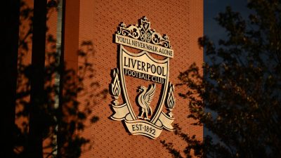 Liverpool FC crest on a red brick wall, illuminated by sunlight with tree shadows.