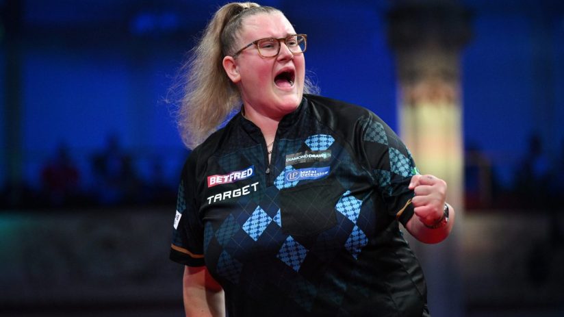 Blonde woman in glasses and a patterned shirt, with her mouth open and fist clenched, celebrating during a darts match