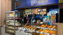 Interior of a Greggs bakery with a staff member behind the counter, displays of pastries, drinks, and The Big Deal signage.