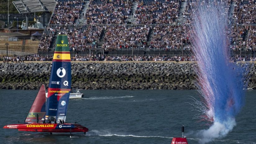SailGP F50 catamaran Los Gallos sailing past a cheering crowd as red and blue smoke erupts.