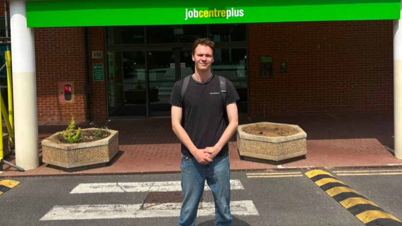 Young man in front of a Jobcentre Plus building, standing on a pedestrian crossing.