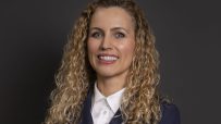 Katie White, a woman with curly blonde hair, smiling in a navy blazer and white collared shirt against a dark background.