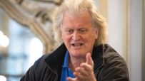 Tim Martin, founder of JD Wetherspoon, speaking and gesturing with an open hand, wearing a blue polo shirt and dark jacket.