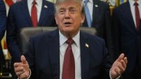 Donald Trump speaking at a desk, gesturing with hands, wearing a dark suit and red tie.