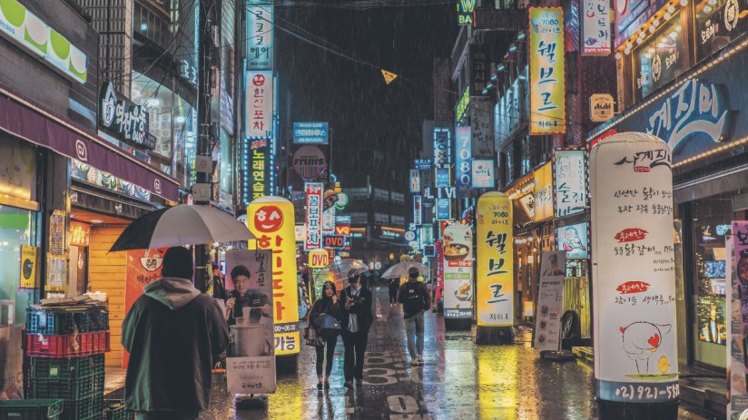 Rainy night street in Seoul, South Korea, with neon signs, storefronts, and people with umbrellas
