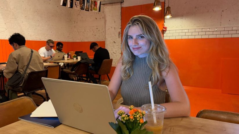 Anna, a young woman, smiles while typing on a laptop at a cafe table with a drink and potted plant.