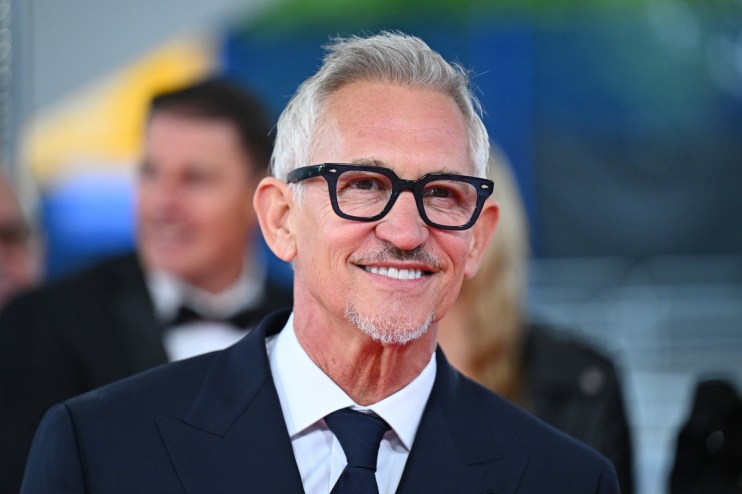 Gary Lineker smiling in a dark suit, white shirt, and navy tie, wearing black-framed glasses.