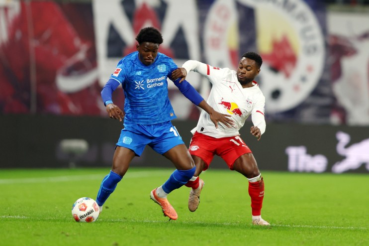 Two male soccer players, one in blue and one in white/red, vie for the ball on a green field.