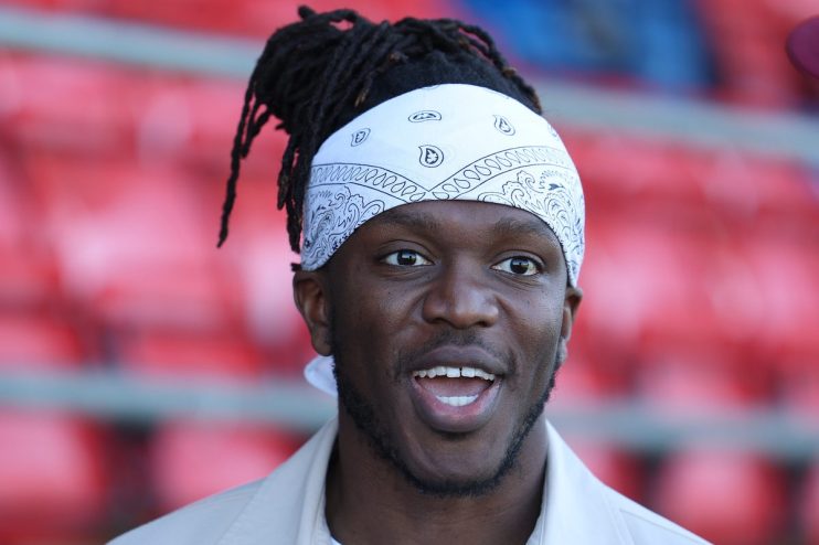 KSI with dreadlocks and a white bandana, smiling with an open mouth, red stadium seats blurred in background.