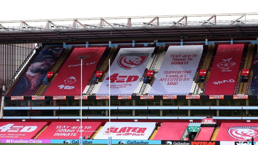 Empty stadium stands with large banners supporting MND awareness, Gloucester Rugby, and the Slater Cup.