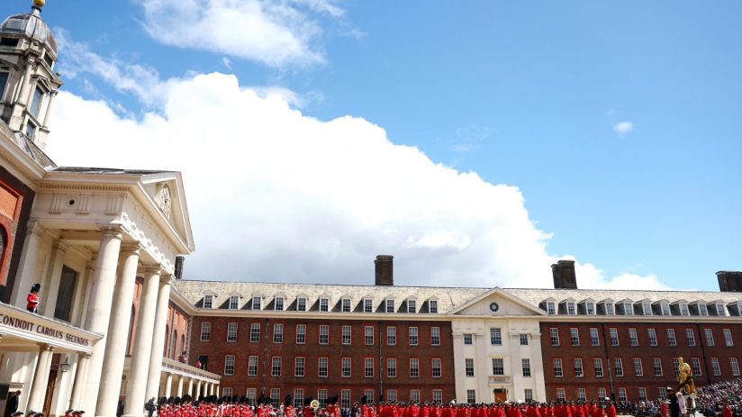 British soldiers in red uniforms parade in front of historic buildings under a partly cloudy sky.