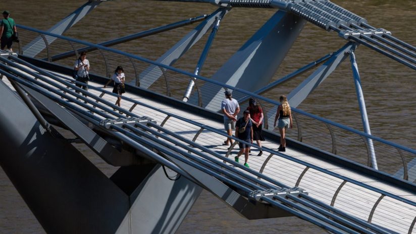 Pedestrians walk across a modern pedestrian bridge with steel cables and supports over brown water.