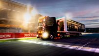 Black Formula 1 hospitality truck driving on a race track at dusk, bright lights and sponsor banners visible.