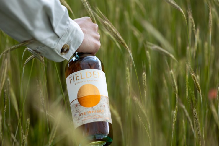 Hand holding Fielden Whisky of England Rye Whisky bottle amidst green rye field.