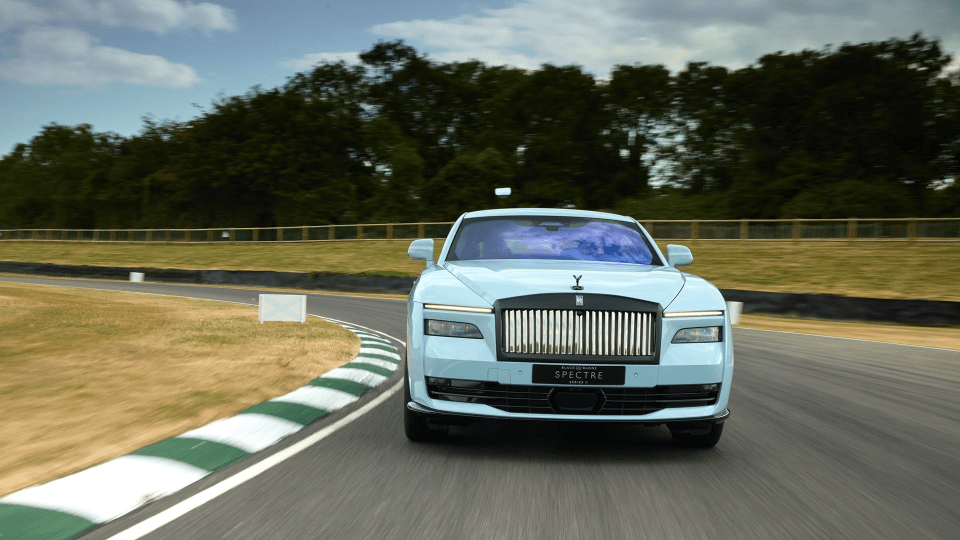 Light blue Rolls-Royce Spectre electric car on a racetrack, with a blurred green landscape in the background.