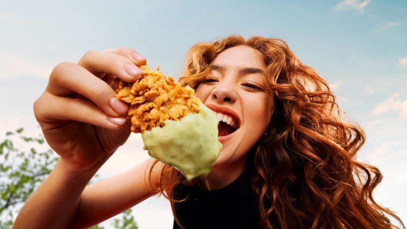 Redhead woman excitedly biting into a piece of KFC fried chicken dipped in green sauce.