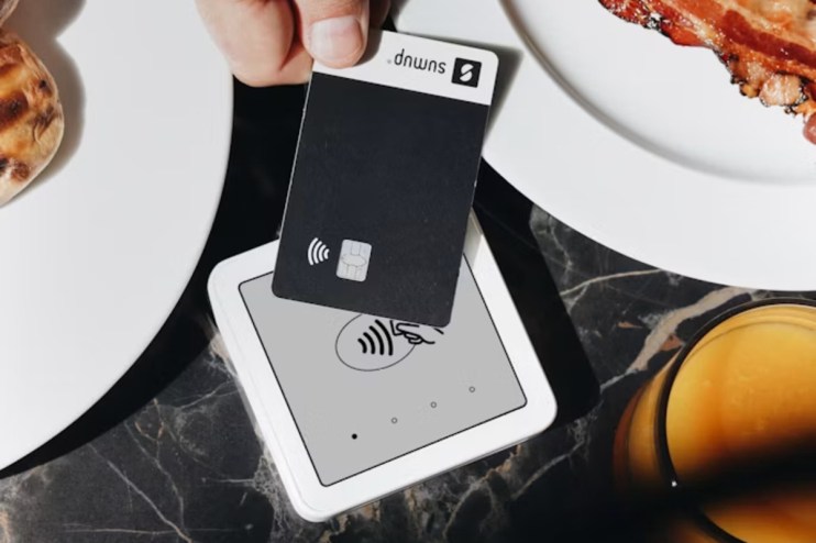 Hand holding black SumUp payment card over a white contactless reader on a marble table with breakfast food