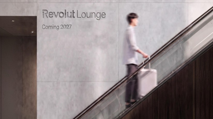 Revolut Lounge sign on a concrete wall with a person on an escalator holding a rolling suitcase.